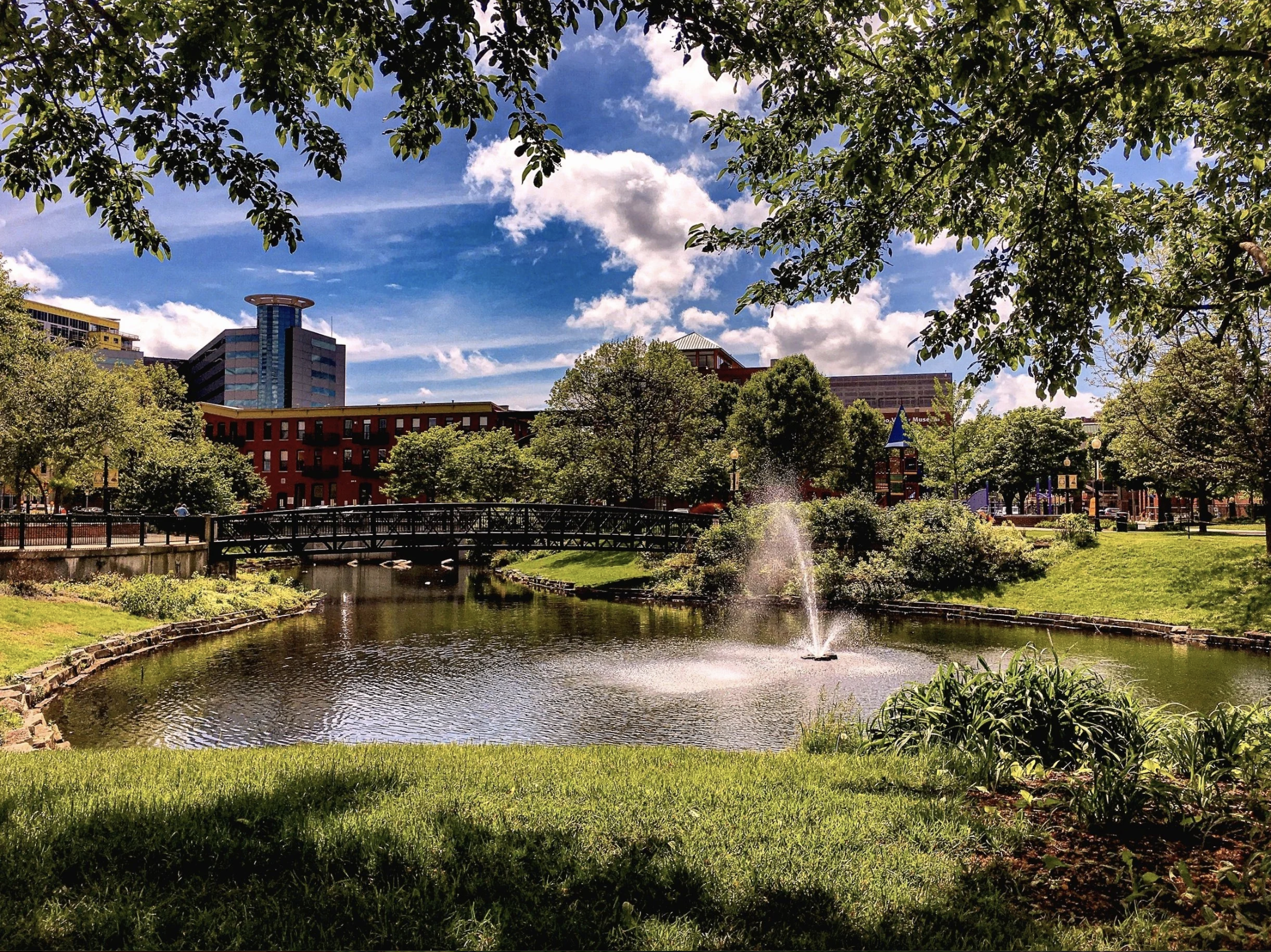 Fountain in a downtown Kalamazoo park with a bridge and city buildings